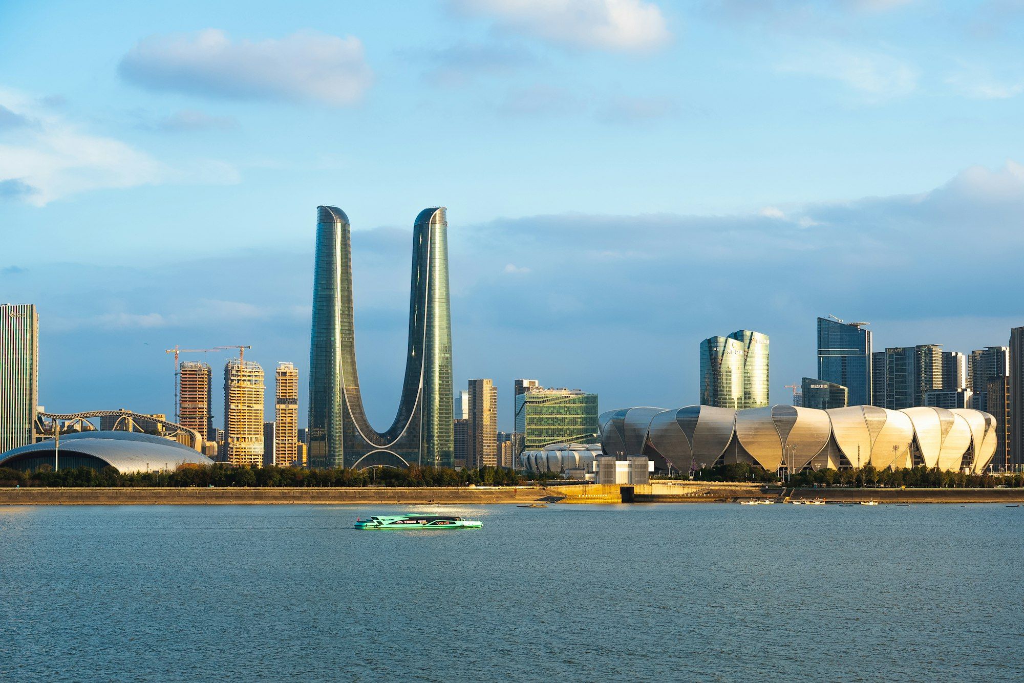 The Hangzhou skyline across the Qiantang River on a clear day, with twin towers joined at the base and a ferry on the water.